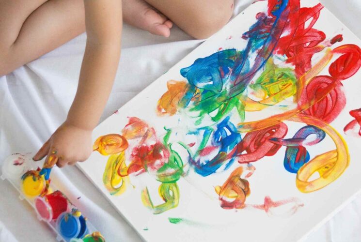 A toddler using finger paint to paint a canvas on a sheet which is a mom hack.