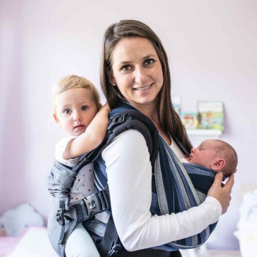 A mom wearing a baby in a ring sling and a second baby on her back in a structured baby carrier.