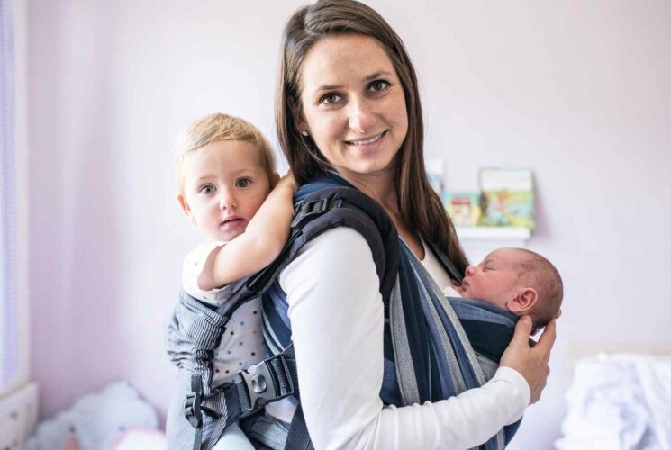 A mom wearing a baby in a ring sling and a second baby on her back in a structured baby carrier.