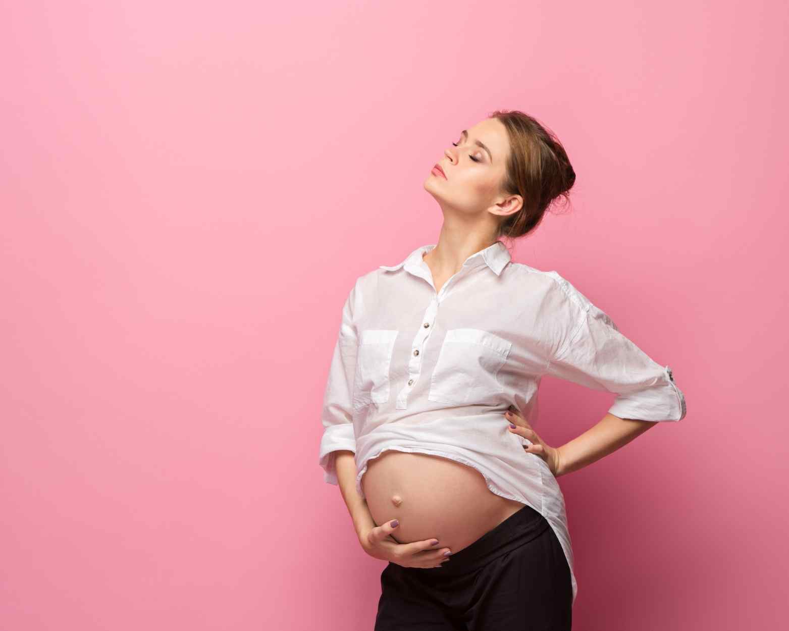 A pregnant woman holding her belly with a pink wall behind her.