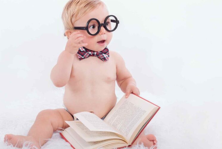 A baby boy sitting in his diaper and holding a book with fake glasses on.