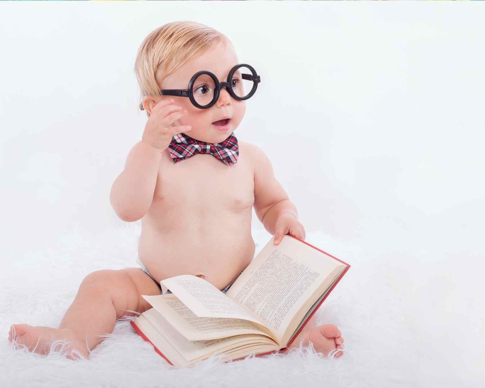 A baby boy sitting in his diaper and holding a book with fake glasses on.