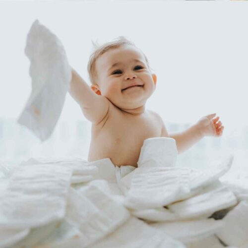 A cute baby boy sitting in a pile of organic diapers and holding up one of the non toxic diapers and smiling.