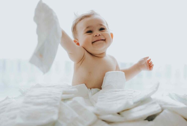 A cute baby boy sitting in a pile of organic diapers and holding up one of the non toxic diapers and smiling.